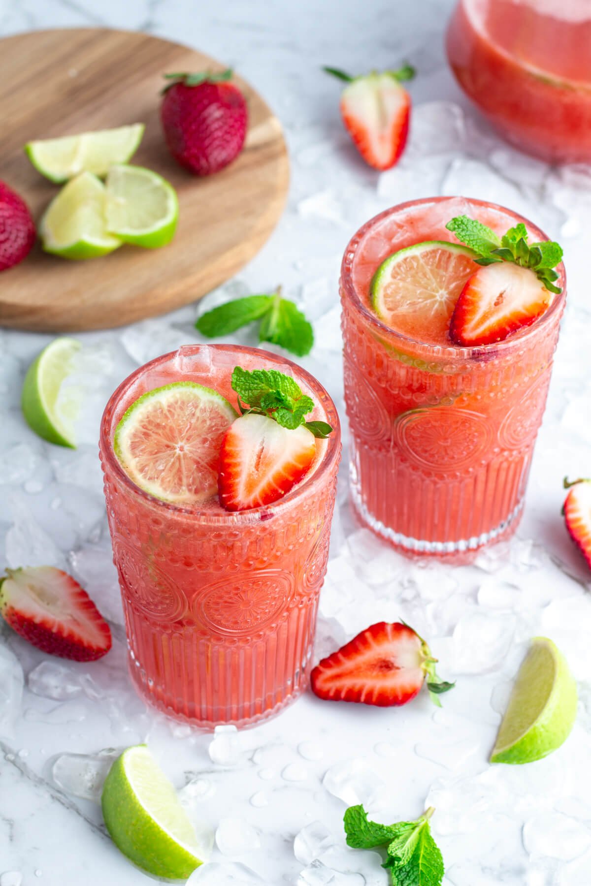 2 tall glasses of strawberry water garnished with strawberry and lime with fresh strawberries, lime wedges and ice scattered around on a white background