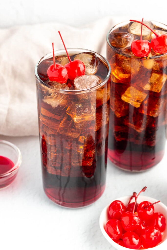 2 tall glasses filled with cola and grenadine and garnished with a maraschino cherry