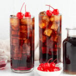 2 Roy Rogers mocktails in tall glasses filled with cola and grenadine with a bowl of maraschino cherries in a bowl