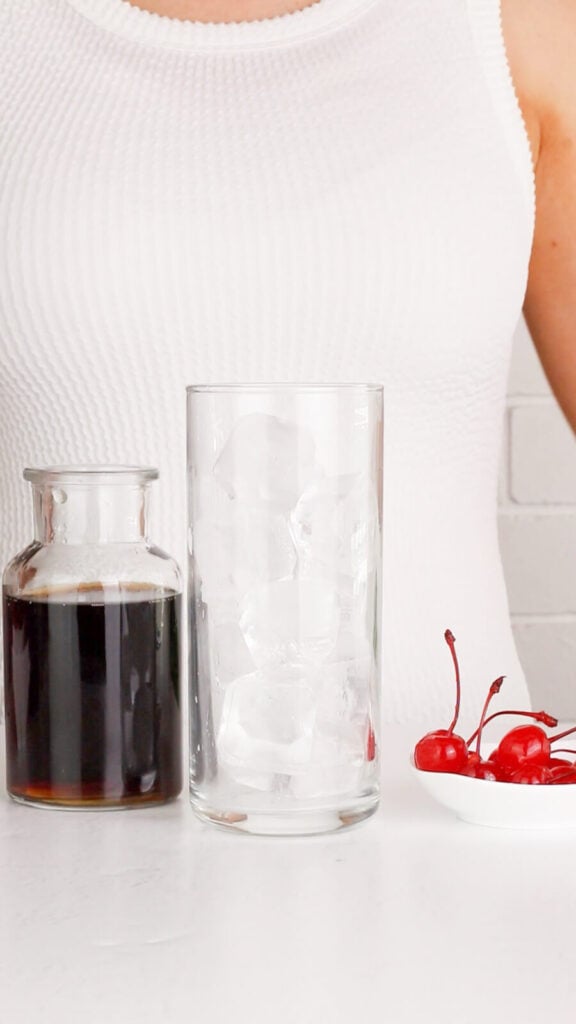 tall glass filled with ice with a bottle of cola to the lift and a bowl of maraschino cherries to the right