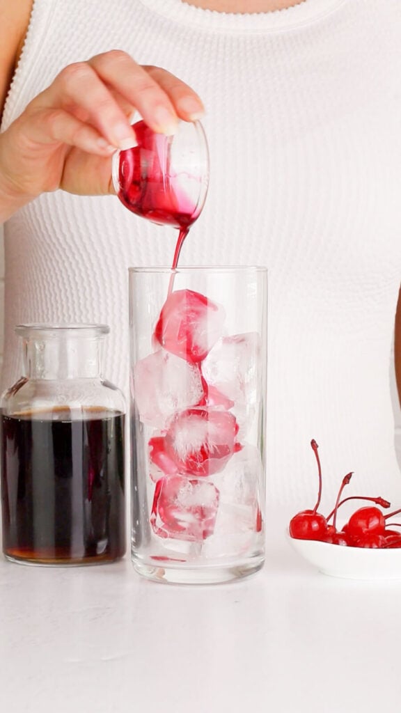 tall ice filled glass with grenadine slowly being poured in on a white background