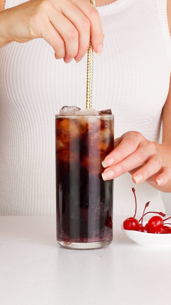 tall glass filled with ice cola and grenadine being stirred with a gold bar spoon