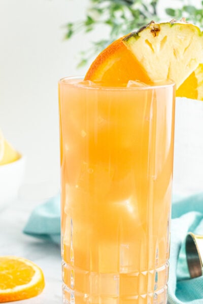 tall glass of orange liquid garnished with an orange slice and pineapple with a blow of lemons in the background