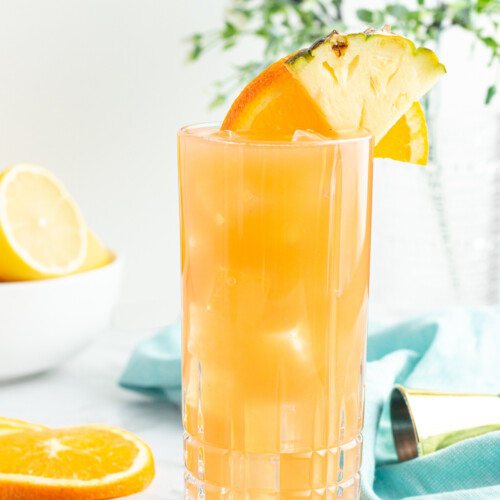 tall glass of orange liquid garnished with an orange slice and pineapple with a blow of lemons in the background
