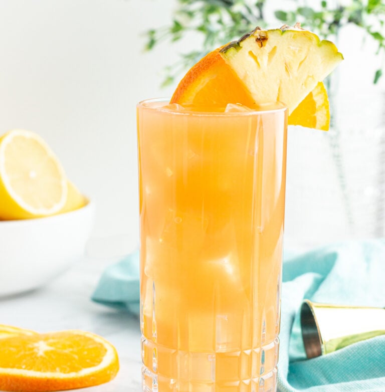 tall glass of orange liquid garnished with an orange slice and pineapple with a blow of lemons in the background