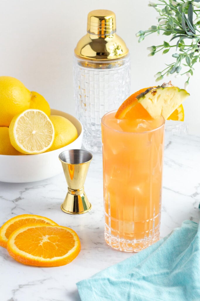 tall glass of orange liquid garnished with an orange slice and pineapple with a blow of lemons in the background