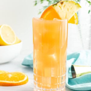 tall glass of orange liquid garnished with an orange slice and pineapple with a blow of lemons in the background