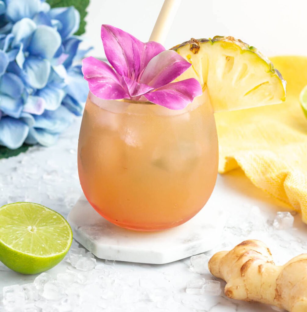 ginger beer mocktail in a pink coloured glass garnished with a purple flower and pineapple slice with a blue flower in the background.