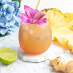 ginger beer mocktail in a pink coloured glass garnished with a purple flower and pineapple slice with a blue flower in the background.