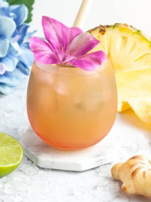 ginger beer mocktail in a pink coloured glass garnished with a purple flower and pineapple slice with a blue flower in the background.