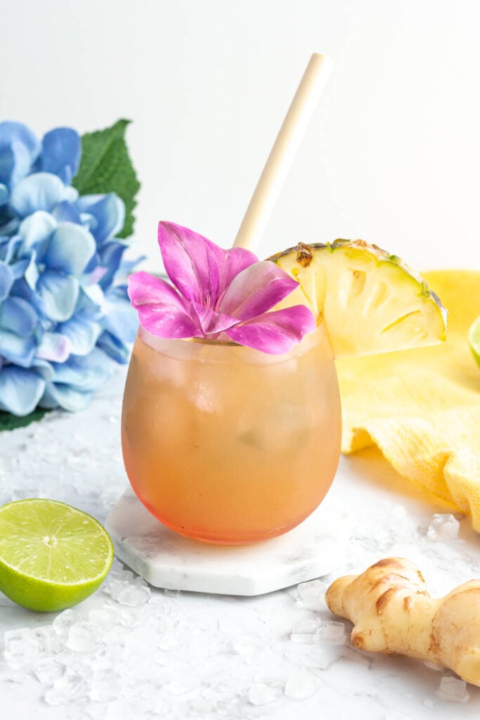 colorful image with a pink drink garnished with a pineapple slice and purple flower and a blue flower in the background.