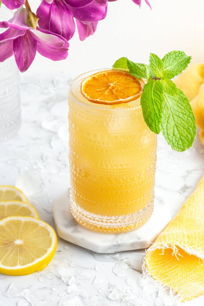 bebida de naranja en un vaso alto adornado con naranja deshidratada y menta con flores de color púrpura en el fondo.
