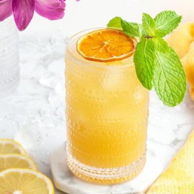 a close up of an orange mocktail in a tall glass with dehydrated orange and mint with purple flowers in the background.