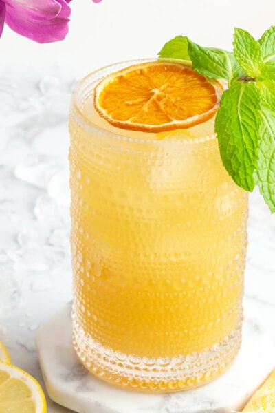 a close up of an orange mocktail in a tall glass with dehydrated orange and mint with purple flowers in the background.