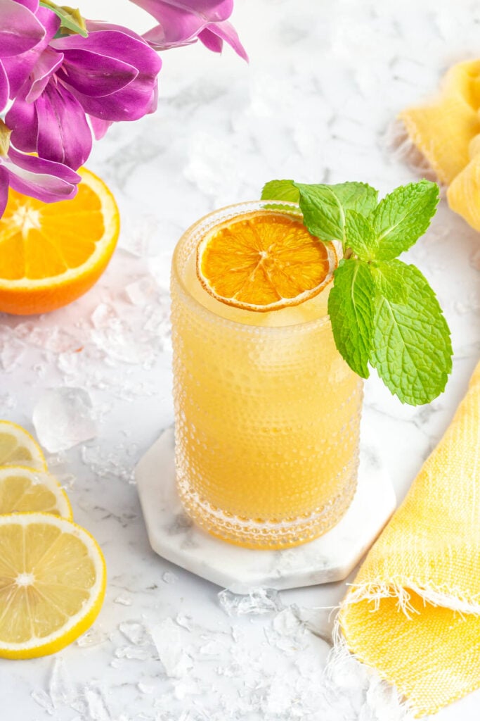 orange drink in a tall glass garnished with dehydrated orange and mint with purple flowers in the background.