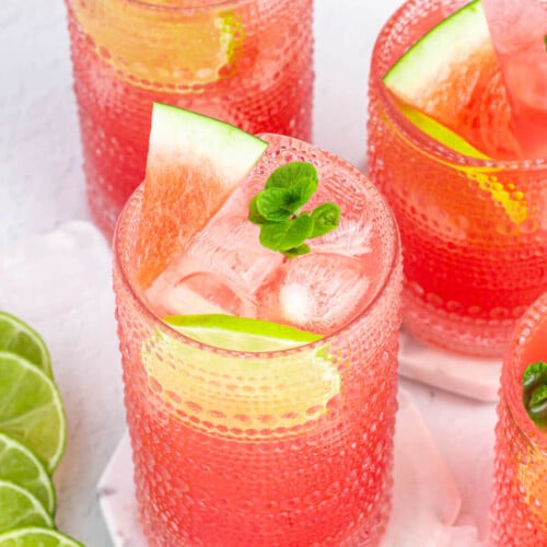 several glass of watermelon fresca garnished with mint, lime and watermelon, with lime rounds in the background.