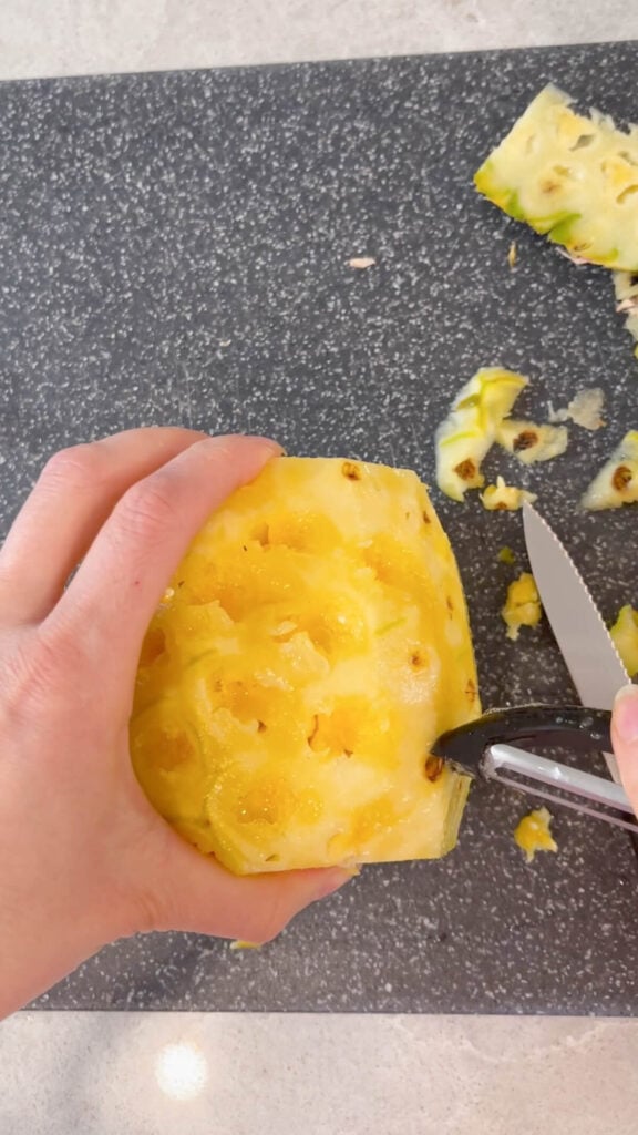 whole peeled pineapple on a black chopping board having the eyes removed by a black vegetable peeler.