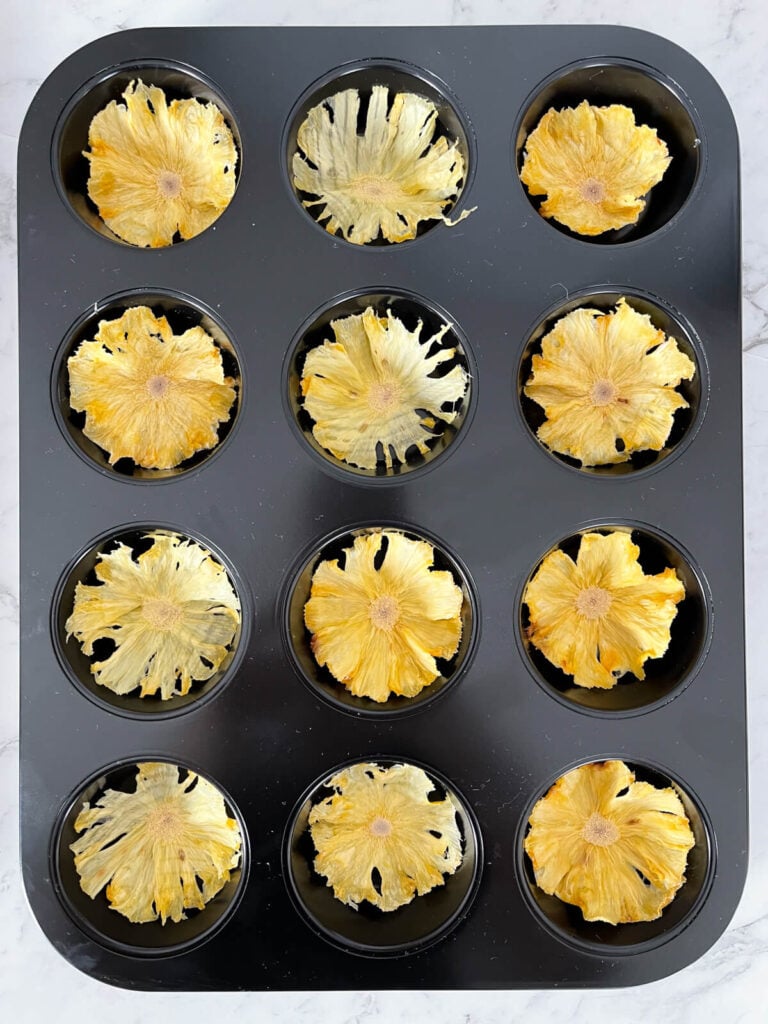 Bandeja negra para mini muffins con una rodaja de piña seca en cada agujero para formar una flor.