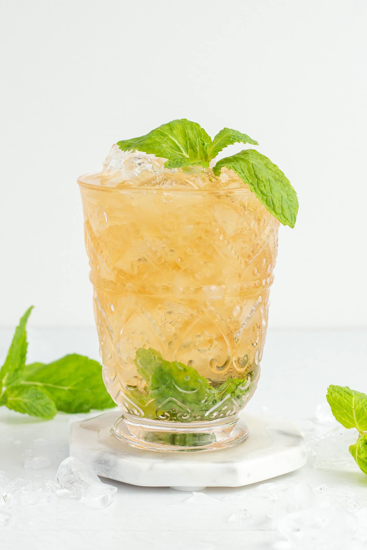 tall glass with intricate pattern with brown liquid, crushed ice and mint inside on a white and grey coaster