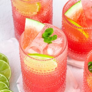 three tall glasses of pink watermelon agua fresca garnished with mint, lime rounds and watermelon wedges.