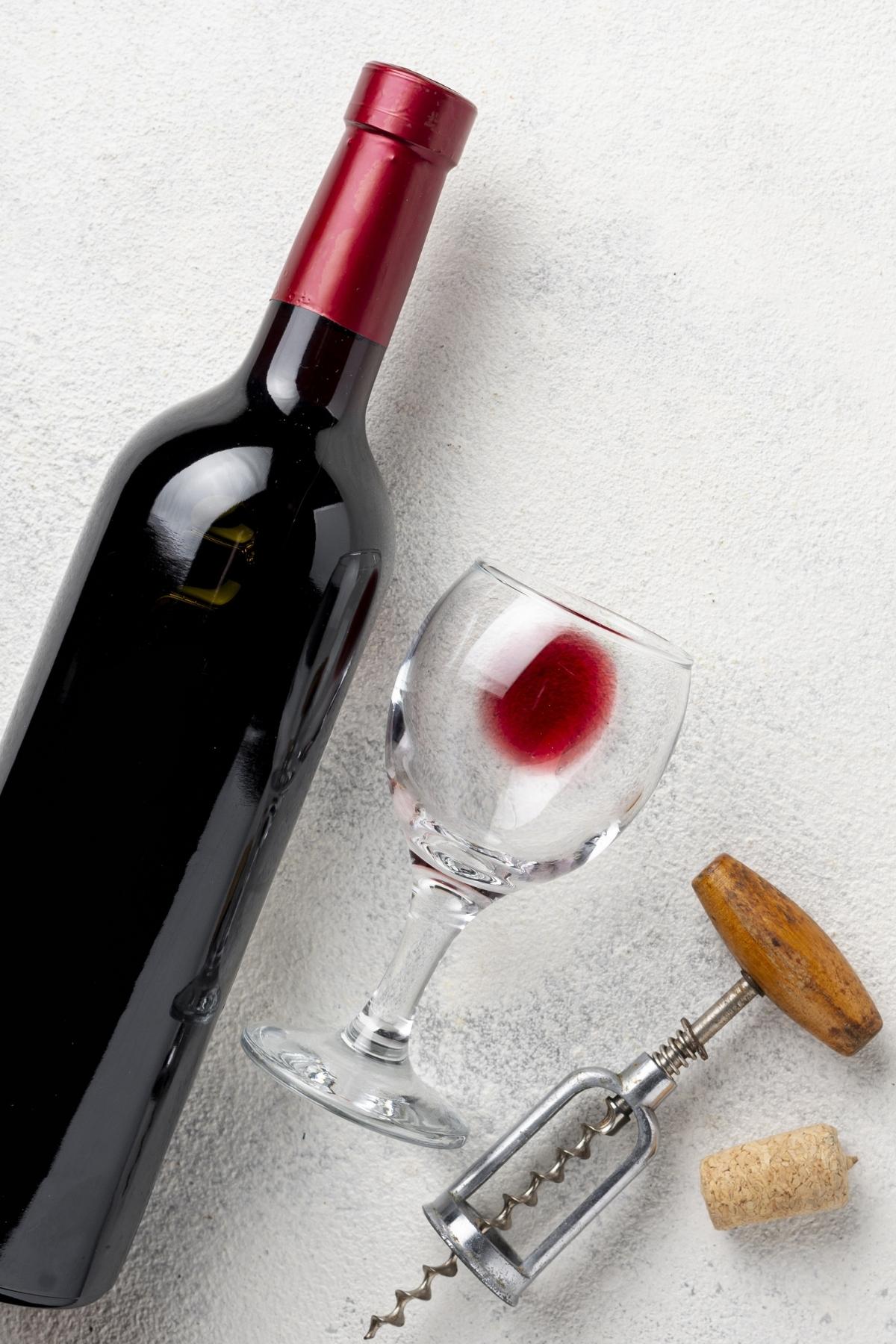 red wine bottle with a glass next to it, and old fashioned bottle opener and a cork.