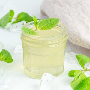 glass jar of mint syrup with fresh mint garnished on top surrounded by a brown tea towel, fresh mint and crushed ice.