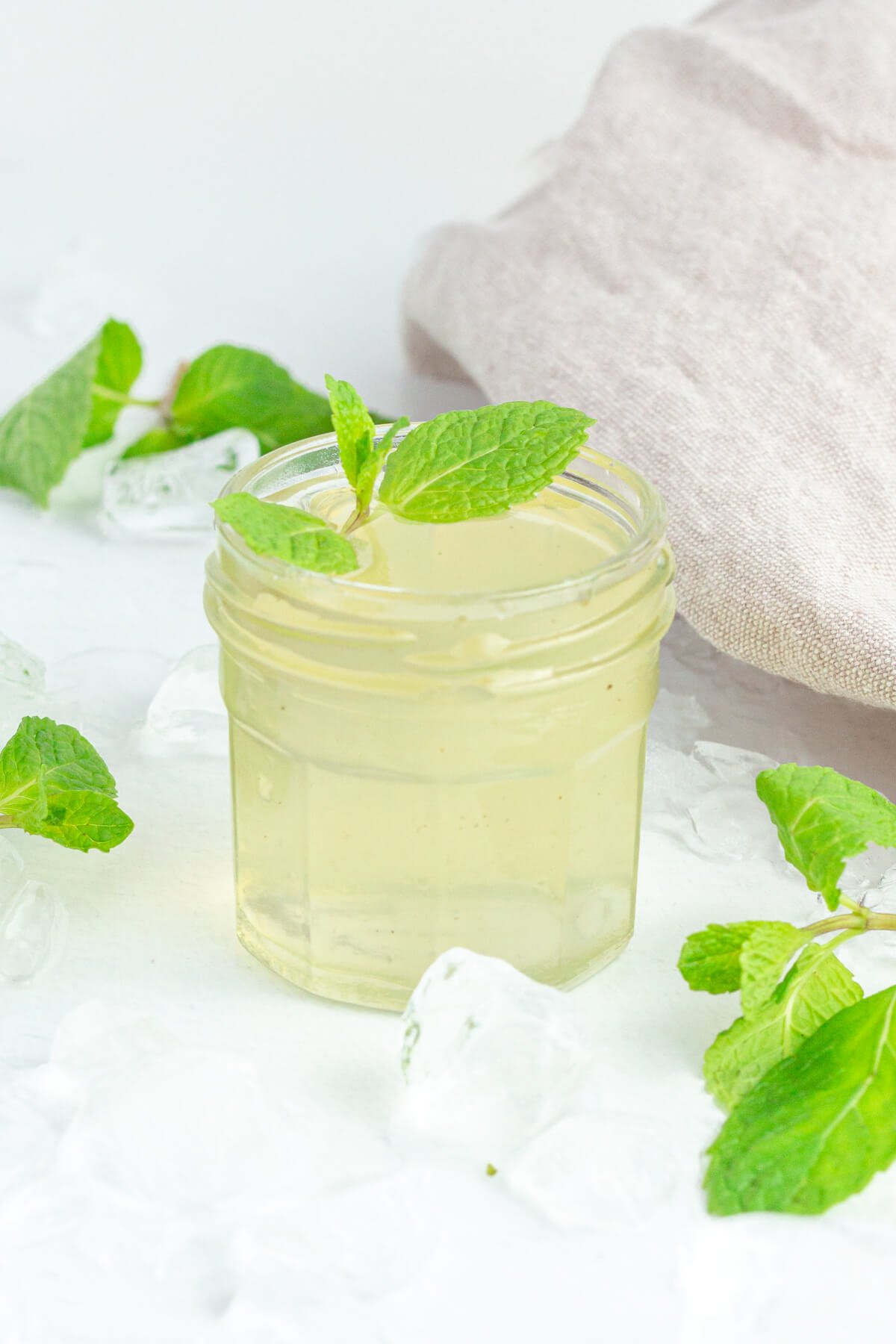 glass jar of mint syrup with fresh mint garnished on top surrounded by a brown tea towel, fresh mint and crushed ice.