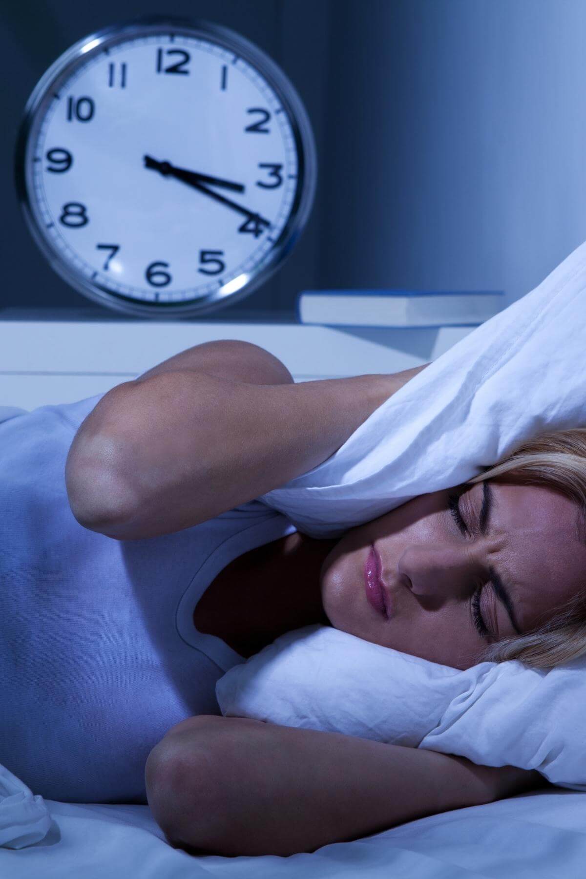 woman lying in bed with a clock behind her, pressing the pillow into her head unable to sleep.