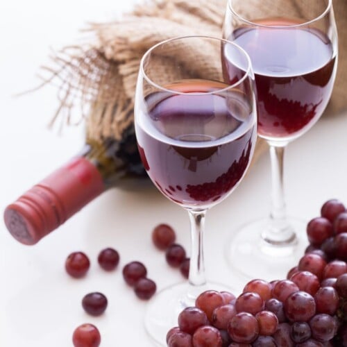 two glasses of alcohol-free red wine surrounded by grapes and a red wine bottle in brown wrapping.