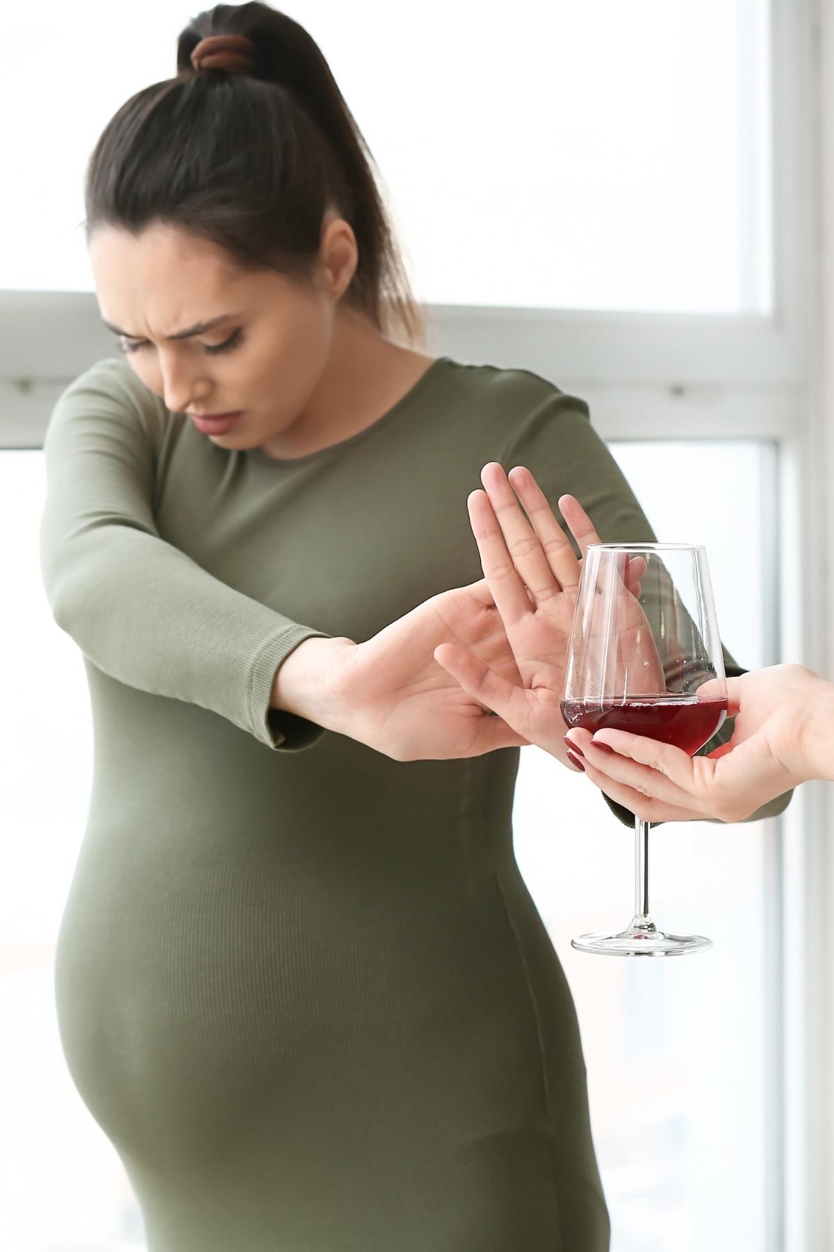 pregnant person in a green top with brown hair pushing away a glass of red wine.