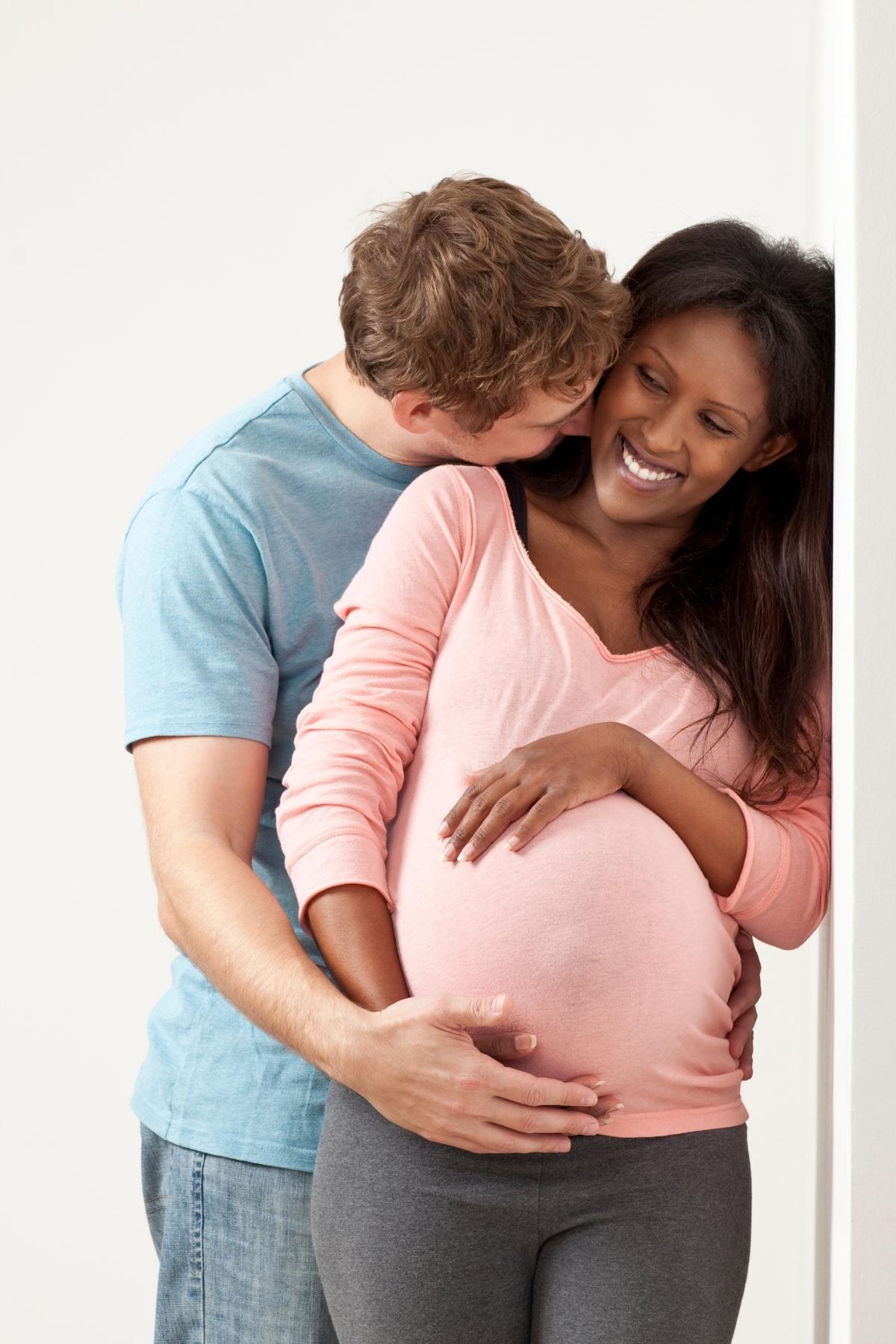 pregnant woman in a pink top and a man in a blue tshirt hugging her from behind.