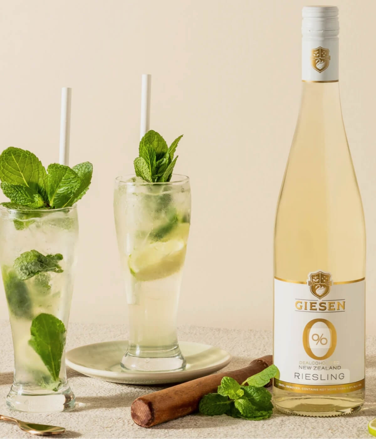 A bottle of Giesen 0% Riesling, one of the best non alcoholic white wines, stands beside two tall glasses filled with ice, mint, lime slices, and straws. A plate with mint leaves and a cinnamon stick rests on the neutral background.