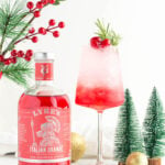 a mocktail with red to white layering in a wine glass garnished with rosemary and cranberries with Christmas ornaments scattered close by, as well as a bottle of Lyre's Italian Orange.