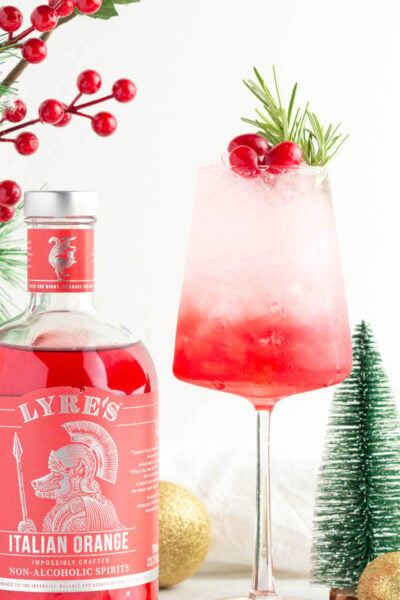 a mocktail with red to white layering in a wine glass garnished with rosemary and cranberries with Christmas ornaments scattered close by, as well as a bottle of Lyre's Italian Orange.