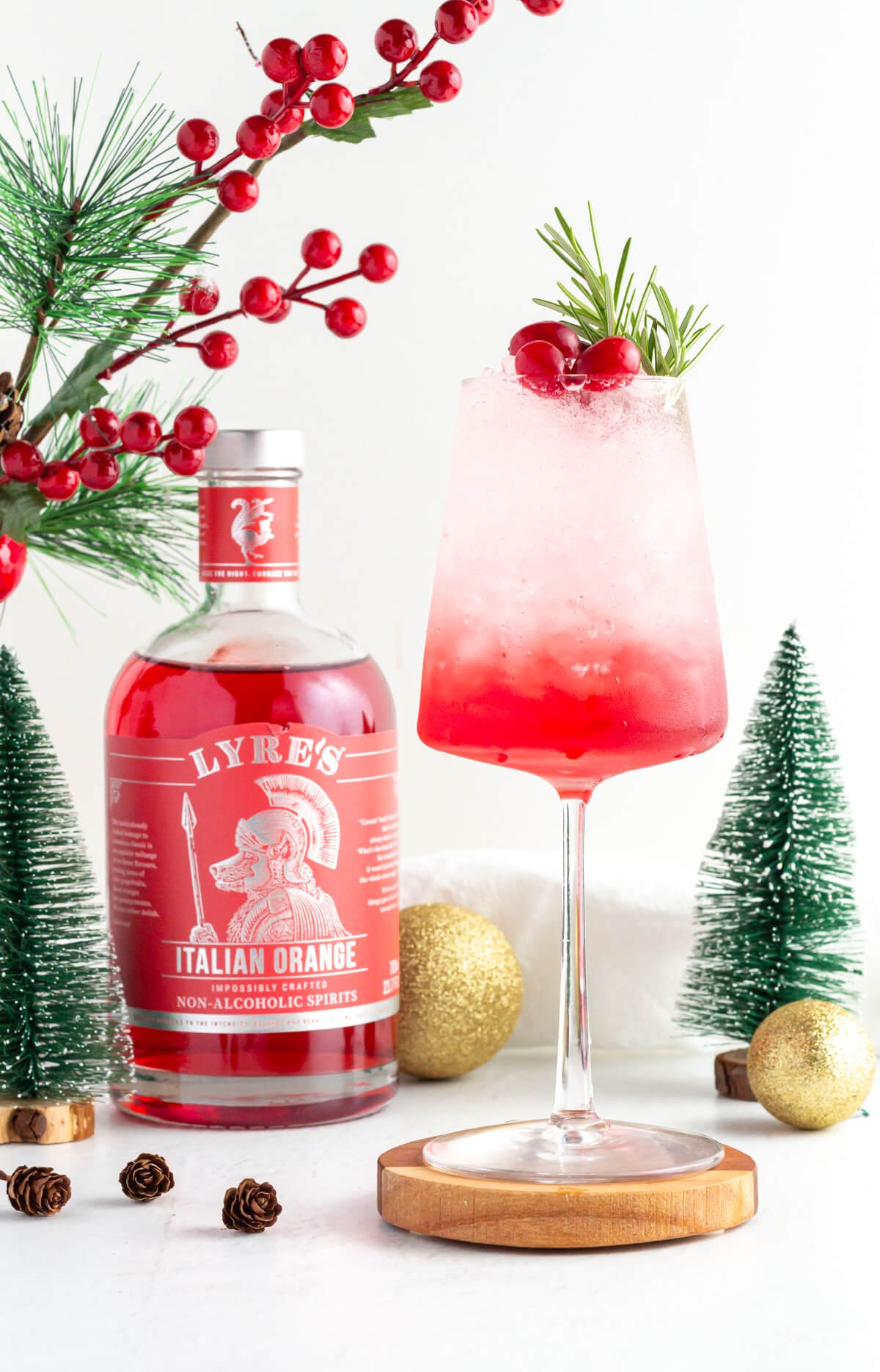 a layered mocktail in a wine glass with red at the bottom and white at the top garnished with rosemary and cranberries with Christmas ornaments scattered close by, as well as a bottle of Lyre's Italian Orange.