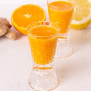 two wellness shots made with ginger turmeric and orange in glass shot glasses on a white background.