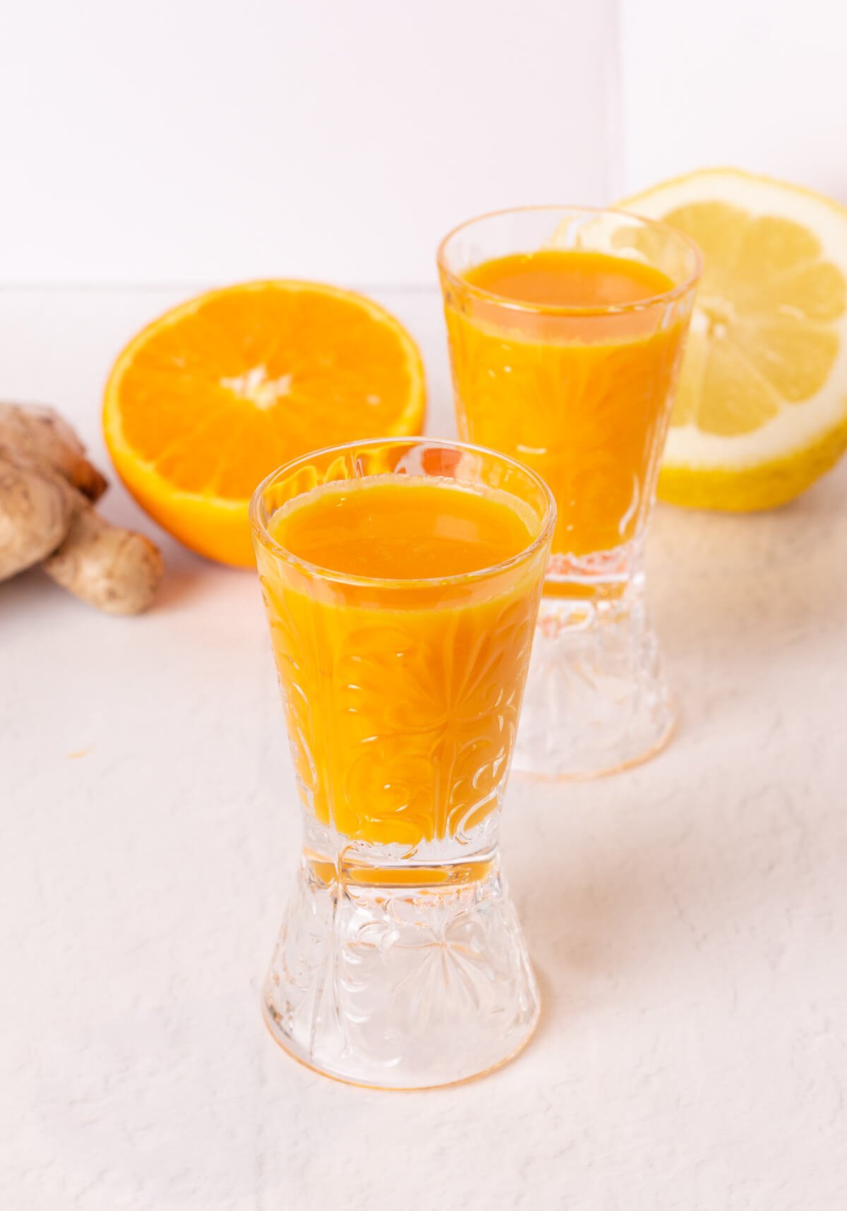 two glass shot glasses filled with an orange liquid of ginger and turmeric with orange, lemon and ginger in the background.