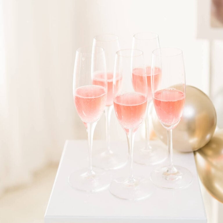 a white table with 5 champagne flutes of non alcoholic rosé wine with gold balloons in the background.
