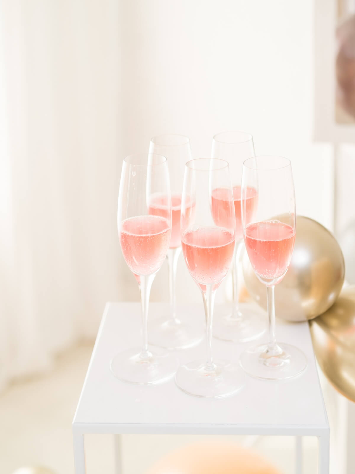 a white table with 5 champagne flutes of non alcoholic rosé wine with gold balloons in the background.