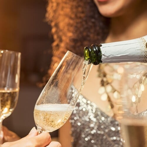 a festive vibe with a champagne bottle pouring sparkling wine into a champagne group with a girl dressed in glitter blurred in the background.