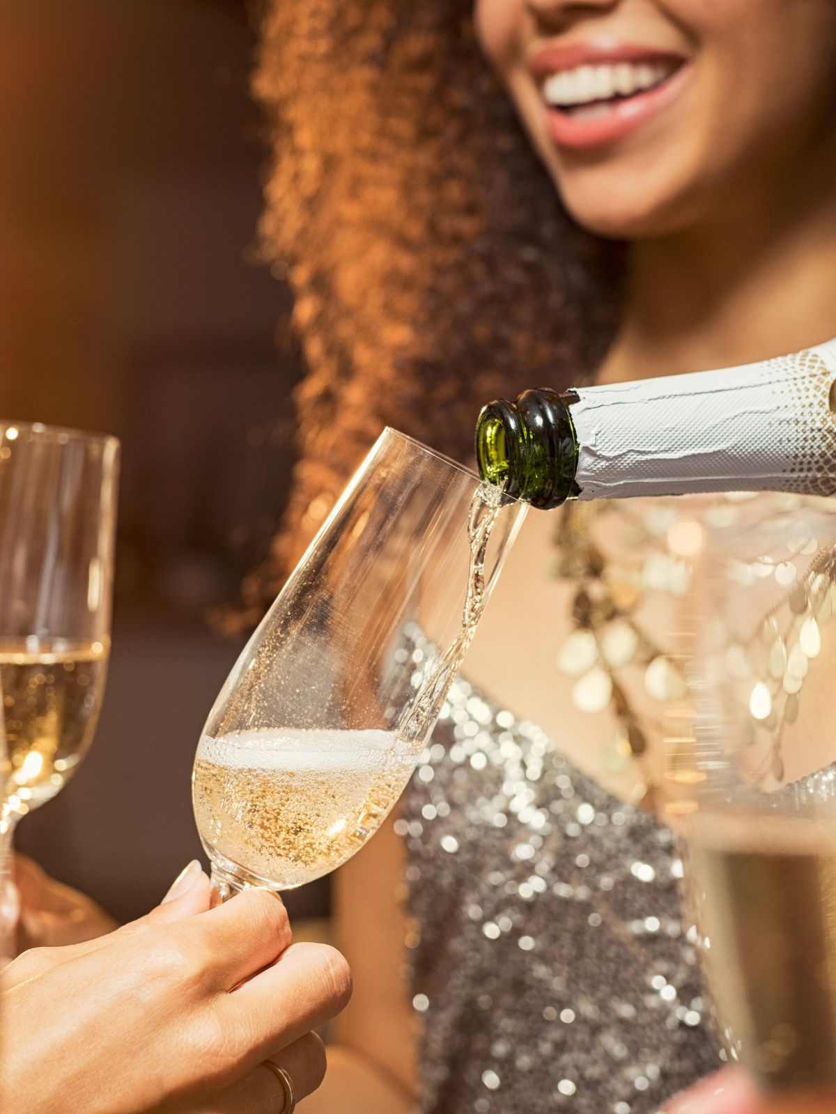 a festive vibe with a champagne bottle pouring sparkling wine into a champagne group with a girl dressed in glitter blurred in the background.