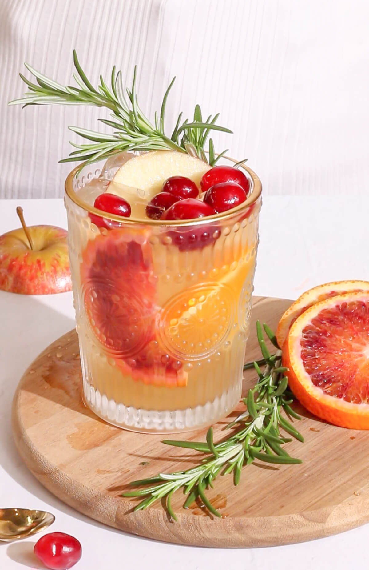 a yellow-orange punch in an intricate glass tumbler with a gold rim garnishe with blood orange cranberries and rosemary on a wooden board with rosemary orange and apple scattered around it.