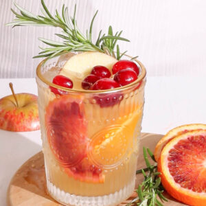 a pretty thanksgiving inspired punch that is light orange in color in a glass tumbler with a gold rim garnished with blood orange and cranberries.