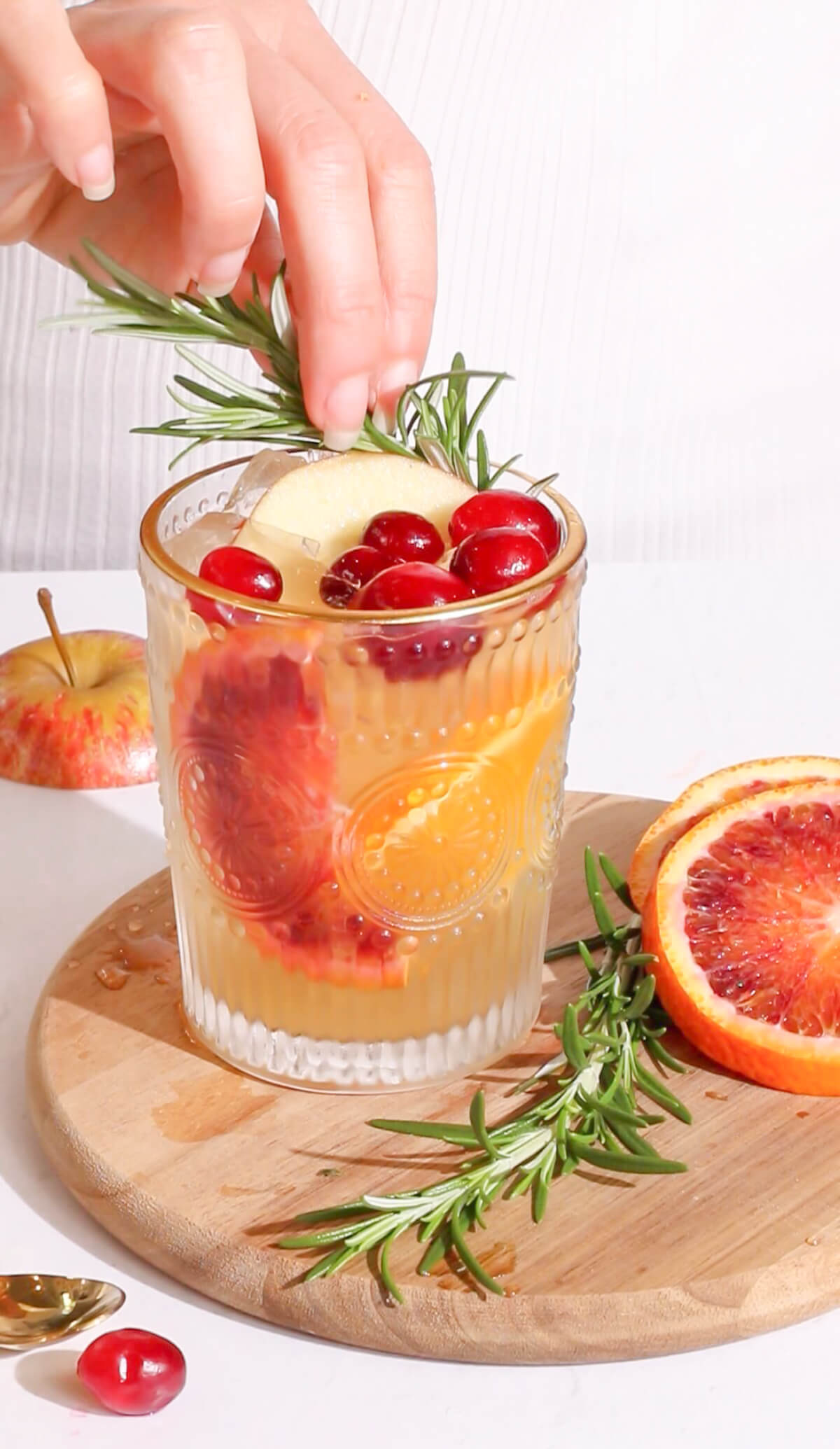 a fruity punch in a intricate glass with a gold rim garnished with rosemary cranberries apples and pear with a hand touching the rosemary garnish.