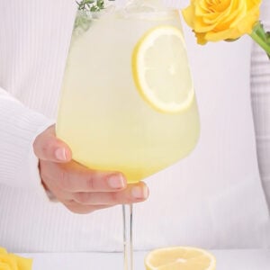 large spritz glass with non-alcoholic limoncello, alcohol-free prosecco and soda water inside, garnished with a slice of lemon and piece of thyme on a white background.