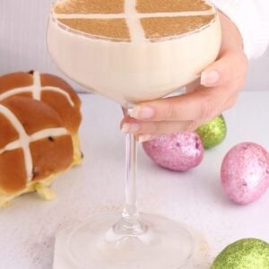 creamy colored mocktail that resembles a hot cross bun in a coupe glass with green and pink easter eggs scattered underneath.