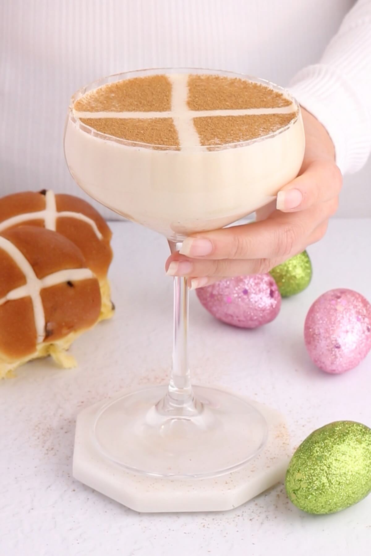 creamy colored mocktail that resembles a hot cross bun in a coupe glass with green and pink easter eggs scattered underneath.