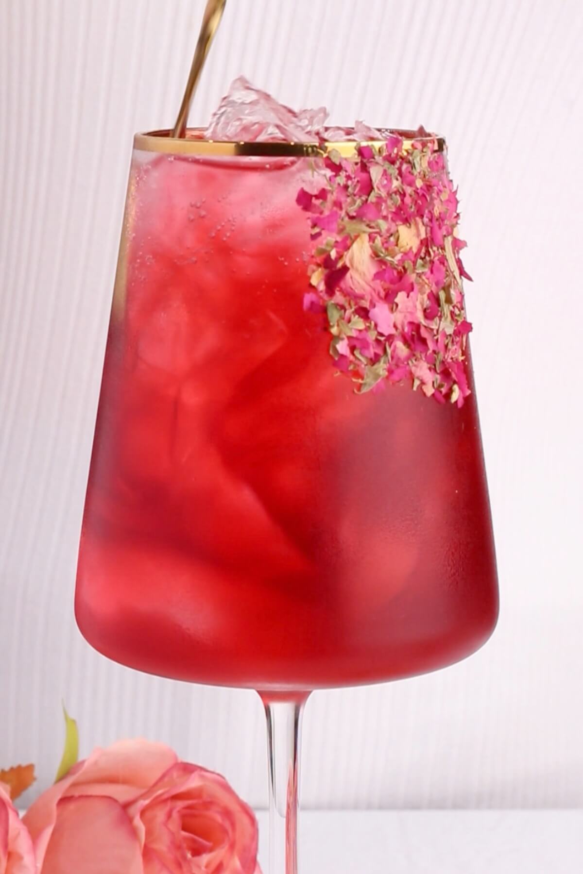 close up of pink mocktail recipe in a wine glass that has a gold rim and edible rose petals stuck on the side.