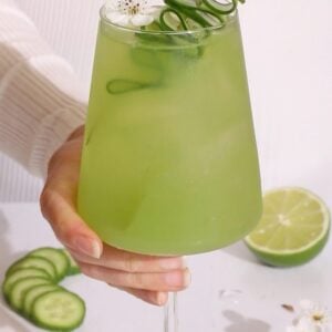 green mocktail with cucumber lime and elderflower in a wine glass garnished with a cucumber ribbon and a white flower.