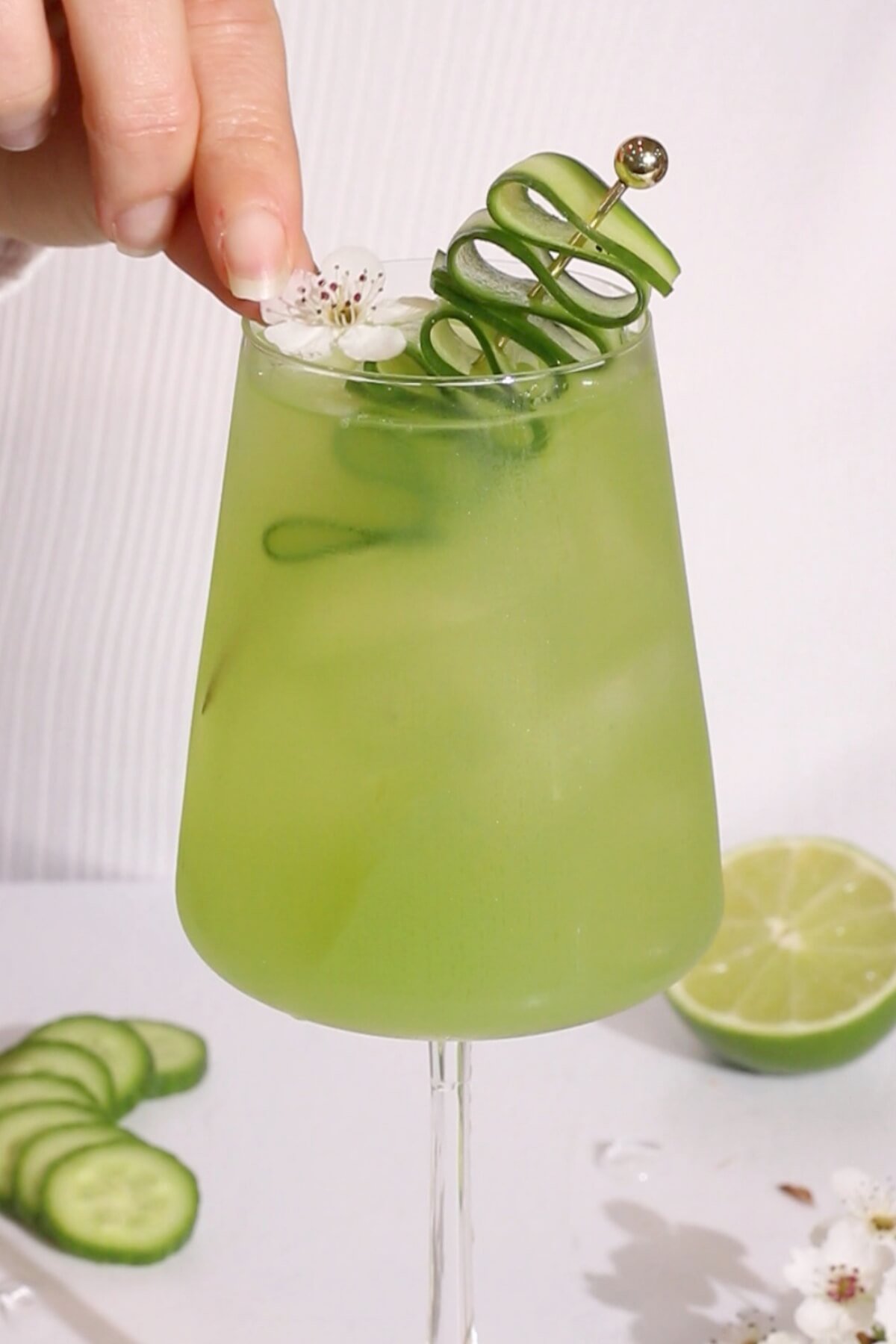 cucumber and lime non alcoholic cocktail in a wine glass garnished with a cucumber ribbon and a white flower.